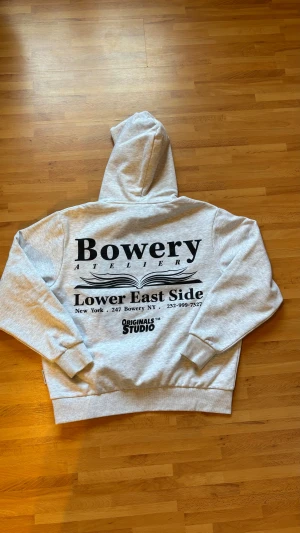 Grå Bowery hoodie med tryck - Snygg ljusgrå hoodie från Jack & Jones (Bowery) med stor svart text och grafiskt tryck på ryggen samt mindre logga på bröstet. Tröjan har huva, känguruficka och ribbade muddar. Perfekt för dig som gillar streetwear och vill ha en chill look.