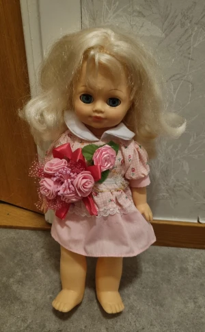 Söt rosa blommig dockklänning - Dockklänning i ljusrosa bomull med små blommor, vit krage och spetsdetaljer. Puffärmar och volangkant nertill ger en gullig look. Klänningen har ett band i midjan och matchas med en bukett rosa tygrosor med röd rosett.