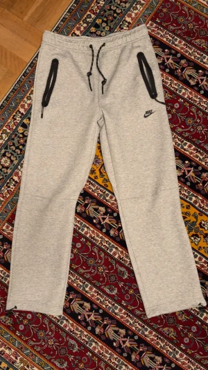 Grå mjukisbyxor från Nike med snörning - Grå mjukisbyxor från Nike med svarta dragkedjor på fickorna, justerbar snörning i midjan och liten Nike-logga på låret. Byxorna har raka ben och dragsko vid benslut för extra passform. Perfekta för chill eller träning.