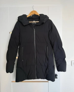 Svart dunjacka med huva från Uniqlo - Svart dunjacka från Uniqlo i storlek S. Jackan har en snygg, lång passform med huva och två praktiska fickor med dragkedja. Materialet är polyester på utsidan och dunfyllning för extra värme. Perfekt för kalla dagar och har en stilren look.
