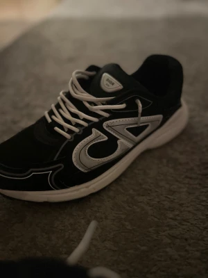 Svarta Dior sneakers med vita detaljer - Säljer ett par svarta Dior sneakers med vita snören i storlek 44 och tydliga vita detaljer på sidorna, inklusive den ikoniska CD-loggan. Skorna har en rund tå, platt sula och är tillverkade i syntetmaterial med inslag av skinn. Perfekt för dig som gillar lyxiga och trendiga sneakers.