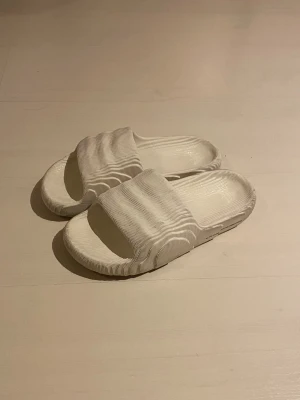 Vita Adidas Adilette 22 - Säljer ett par vita sandaler med ribbad struktur och öppen tå. De har en bred rem över foten och är tillverkade i syntetmaterial. Sulan är platt och mönstrad för extra grepp. Perfekta för chill dagar och sommarhäng.