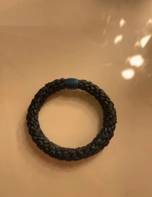 Svart flätat armband från Kknekki - Snyggt svart flätat armband från Kknekki med en rund blå plastdetalj där märket är ingraverat. Armbandet har en grov och glansig struktur som ger en cool och modern vibe. Perfekt att bära som en statement-accessoar.