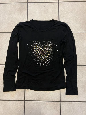 Svart långärmad tröja med hjärtmotiv storlek S/M - Svart långärmad tröja med rund hals och stort hjärtmotiv framtill, dekorerat med pärlor i guld, silver och vitt. Mjuk och tunn i materialet, perfekt för dig som gillar detaljer och vill sticka ut lite extra. Snygg att matcha med jeans eller kjol.