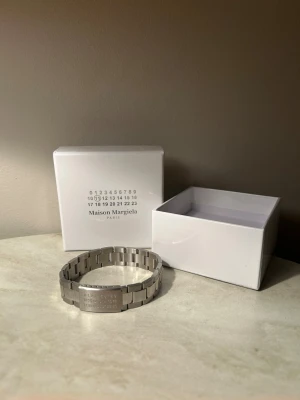 Maison Margiela silverfärgat armband - Snyggt armband från Maison Margiela i silverfärgat rostfritt stål. Länkarna är breda och har en modern, chunky design. Plattan framtill har Margielas ikoniska siffror ingraverade. Kommer med original vit box.