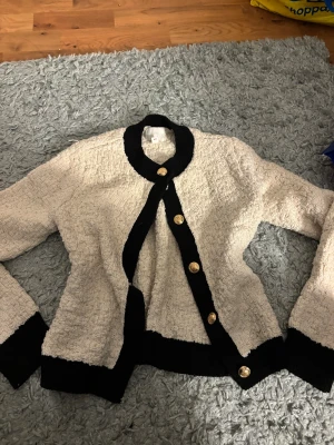 Beige kofta med svarta detaljer från H&M - Snygg beige kofta från H&M med svarta ribbade kanter och guldiga knappar framtill. Koftan har långärmad design och är tillverkad i ett mjukt, strukturerat material som ger en cool vintagekänsla. Perfekt för dig som gillar klassisk stil med en twist.🤍🤍