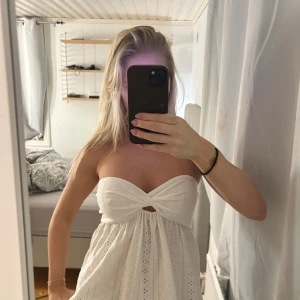 Vit axelbandslös topp - Säljer en söt vit axelbandslös topp med broderade detaljer och ett twistat parti framtill som skapar en liten öppning. Klänningen har ett luftigt fall och är perfekt för varma dagar. Materialet känns som bomull och har ett fint hålmönster.