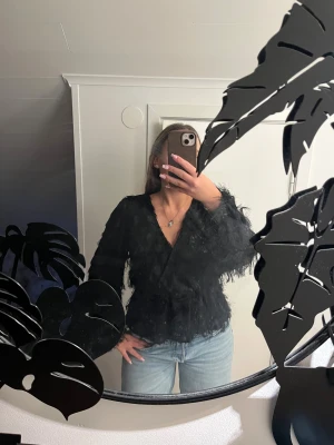 Svart fluffig omlottopp med v-ringning - Säljer en svart omlottopp med v-ringning och långa ärmar.💓 Toppen har en fluffig, fjäderliknande textur, passar till såväl middag som fest🫶🏼