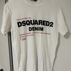Vit DSQUARED2 t-shirt med tryck - Säljer en vit t-shirt från DSQUARED2 med stort svart och rött tryck på bröstet. Modellen är klassisk med rund hals och korta ärmar. Tillverkad i mjuk bomull och har en clean vibe med cool text framtill.  Storlek M