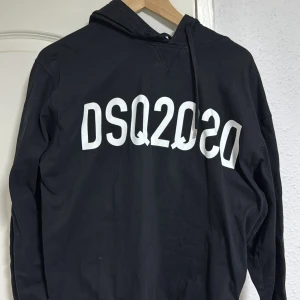 Svart DSQUARED2 hoodie med tryck - Svart hoodie från DSQUARED2 i storlek L med stor vit DSQ2DSQ-logga på bröstet. Tröjan har huva med snörning och är tillverkad i mjuk bomull. Storlek L men passar M 