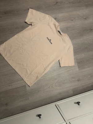 Beige Stone Island t-shirt med tryck - Snygg beige t-shirt från Stone Island med svart logotyptryck på bröstet och stort, slitet logotryck på ryggen. Klassisk rund hals och korta ärmar. Perfekt för dig som gillar streetwear och vill ha något stilrent med edge.