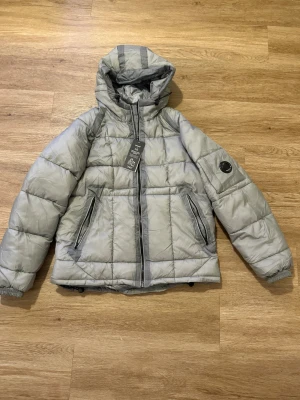 Grå pufferjacka från C.P. Company - Grå pufferjacka från C.P. Company med huva och ikonisk linsdetalj på ärmen. Jackan har två snedställda fickor med dragkedja, quiltad design och elastiska muddar. Materialet är polyester och jackan har en snygg, modern look som passar perfekt till kyliga dagar.