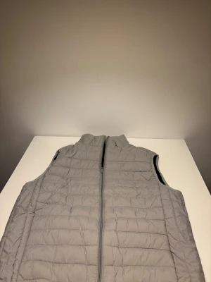 Ljusgrå quiltad dunväst  - Quiltad dunväst i ljusgrå färg med hel dragkedja framtill och ståkrage. Västen har ett stilrent, rakt snitt och horisontella sömmar som ger en modern look. Perfekt att slänga över en hoodie eller tröja för extra värme under kyliga dagar. Helt ny.
