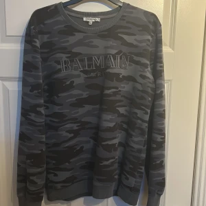 Grå camouflagemönstrad tröja från Balmain - Säljer en snygg grå camouflagemönstrad tröja från Balmain. Tröjan har en rund halsringning och långa ärmar. Perfekt för en stilren och modern look.