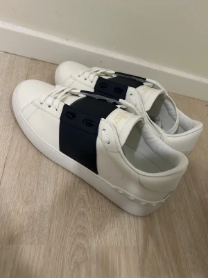 Valentino Garavani Open sneakers vit/blå - Snygga Valentino Garavani Open sneakers i vitt skinn med bred marinblå rem över snörningen. Platt sula och rund tå, samt ikoniska nitar baktill på hälen. Guldtext på tungan och vita skosnören. Perfekt för dig som gillar exklusiv streetstyle. Helt nya allt följer med paketet 