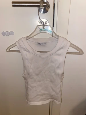 Vit ribbad linnetopp från Zara - Säljer en vit ribbad linnetopp från Zara i storlek S. Toppen är ärmlös med klassisk rund halsringning och har en tight passform. Perfekt att styla med jeans eller kjol för en clean look.