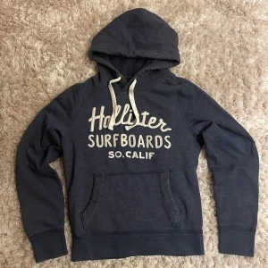 Mörkblå Hollister hoodie med tryck - Snygg mörkblå hoodie från Hollister med vit text 'Surfboards SO.CALIF' på bröstet. Riktigt schysst hoodie i storleken S. Sitter perfekt på dig som har storleken S.