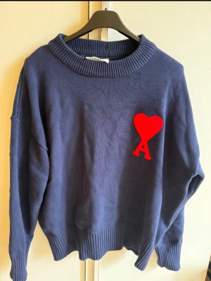 Marinblå stickad tröja från Ami - Säljer en marinblå stickad tröja från Ami med ett stort rött hjärta och bokstaven A broderat på bröstet. Tröjan har ribbad krage, muddar och nederkant samt långärmad passform. Perfekt för dig som gillar stilrena detaljer och vill sticka ut med en ikonisk design.