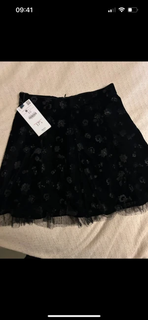  Ny Svart  kortkjol i sammet  - Supersnygg svart kortkjol från Bershka med blommigt mönster och transparent tyllkant längst ner. Kjolen är gjord i mjuk sammet och har en klassisk A-linjeform som ger en cool och trendig vibe.