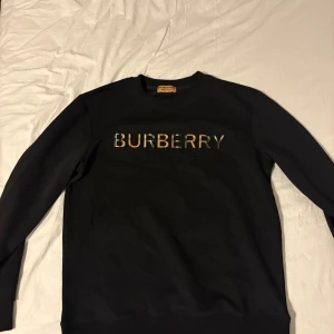 Svart Burberry sweatshirt med logga - Svart sweatshirt från Burberry med klassisk rund hals och stort Burberry-tryck i rutigt mönster på bröstet. Tröjan är tillverkad i mjuk bomull och har långa ärmar. Perfekt för dig som gillar stilrena designer med lyxig touch.
