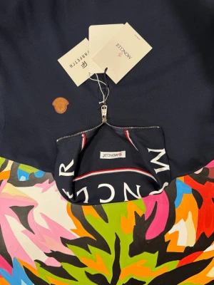 Marinblå Moncler half zip tröja - Snygg marinblå tröja från Moncler med half zip och hög krage där Moncler-loggan är tryckt i vitt. Tröjan har ett brunt Moncler-märke på bröstet och diskreta röda och vita detaljer vid dragkedjan. Perfekt för dig som gillar stilrena och exklusiva plagg.