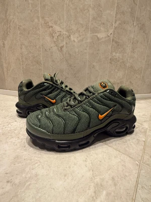 Nike Air Max TN gröna sneakers - Nike Air Max TN sneakers i olivgrönt med orange swoosh och detaljer. Ovandel i mesh och syntet, chunky svart sula med synliga Air-enheter och mönstrad yttersula. Snyggt TN-märke på plösen och sidopaneler med vågiga linjer. Perfekt för dig som gillar streetwear. Är lite små i storleken därför säljer jag.  Aldrig använda utomhus. Har tyvärr ingen box eller kvitto men qr koden leder direkt till Nikes hemsida och köpta i jd butiken i MOS. 