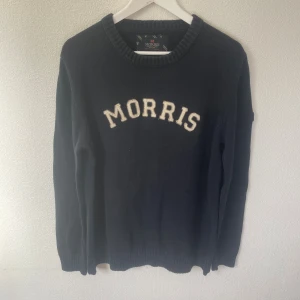 Mörkblå stickad tröja Morris L - Snygg mörkblå stickad tröja från Morris i storlek Large. Tröjan har krämfärgad MORRIS-text framtill och en patch med brittiska flaggan på ärmen. Rund hals och ribbade muddar vid ärmslut och nederkant. Perfekt för dig som gillar klassisk stil.