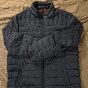 Mörkblå pufferjacka Tommy Hilfiger - Snygg mörkblå pufferjacka från Tommy Hilfiger i storlek M med quiltad design och hög krage. Jackan har dragkedja framtill, två sidofickor med knappstängning och diskret logga på bröstet. Insidan är fodrad i brunt och materialet är polyester. Den högra fickan saknar en knapp så går ej att stänga den, samt så är det en lagning i den vänstra ärmen. Men bortsätt från det så är jackan i bra skick.