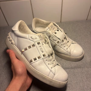 Valentino Rockstud skor - Säljer ett par vita Valentino Rockstud sneakers i skinn med ikoniska silverfärgade nitar. Klassisk rund tå, platt sula och snörning framtill. Märket syns både på plösen och sulan. Riktigt snygga och trendiga skor för dig som gillar lyxiga detaljer.