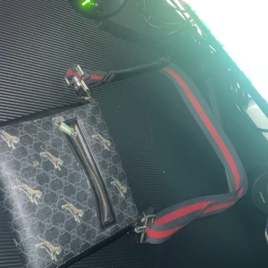  svart gg  axelväska med tigerprint - Svart axelväska från gg med klassiskt GG-mönster och små tigrar över hela väskan. Väskan har en bred axelrem i grått och rött, samt en praktisk dragkedja framtill. Materialet är skinn och insidan är fodrad i svart tyg. Perfekt för dig som vill sticka ut.