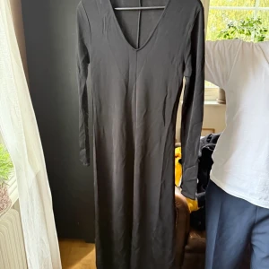 Svart långklänning från Filippa K - Stilren svart långklänning från Filippa K med djup v-ringning och långa ärmar. Klänningen har en smickrande, figurnära passform och är tillverkad i ett mjukt, stretchigt material som känns bekvämt mot huden. Perfekt för dig som gillar minimalistisk och tidlös stil.