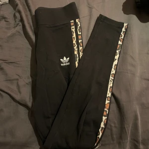 Svarta Adidas leggings med leopardmönster - Snygga svarta leggings från Adidas med klassisk logga på benet och leopardmönstrade ränder längs sidorna. Stretchigt material som sitter tight och bekvämt. Perfekta för dig som gillar sportig stil med en twist.