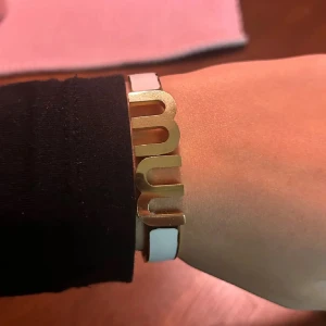 armband i guld och vitt - Snyggt armband med bred, vit bård och stora bokstäver i guldmetall framtill. Armbandet har en stilren och modern design som sticker ut med sin unika form och färgkombination. Perfekt accessoar för att lyfta din outfit.
