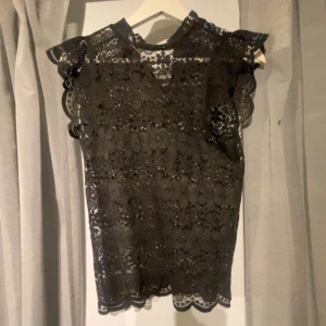 Svart spetsblus med volang - Säljer en svart blus i spets med fina volangdetaljer vid axlarna och ärmlös design. Blusen har ett elegant mönster och en rund halsringning. Perfekt för dig som gillar romantisk och stilren look.