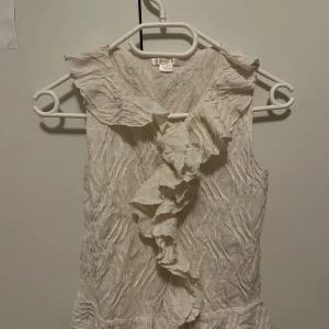 Vit blus med volangdetaljer - använt 2-3 gånger. JAG TAR GÄRNA EMOT PRISFÖRLSAG!!! Säljer en vit blus från Gina tricot med snygga volangdetaljer längs kragen och framtill i storlek 158/164 men passar tilll xxs/xs. Blusen är ärmlös och har ett krinklat tyg med strukturmönster. Perfekt för dig som gillar romantisk och feminin stil.