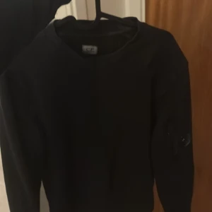 Svart sweatshirt från C.P. Company - Svart sweatshirt från C.P. Company med klassisk rund hals och lång ärm. På vänster ärm finns den ikoniska linsen och en ficka med dragkedja. Tröjan har en clean look och är perfekt för dig som gillar streetwear.