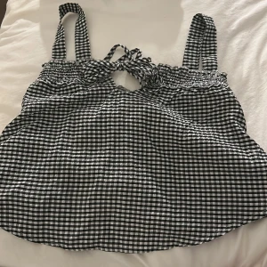 Rutig svartvit linnetopp med knyt - Söt linnetopp i svartvitt rutigt mönster med breda axelband och knytning framtill. Toppen har smock upptill och är luftig och perfekt för varma dagar. Passar till både jeans och kjol. Sååå söt och perfekt för en middag eller utgång!