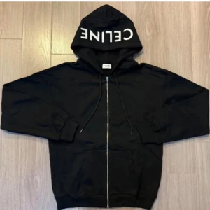 Svart hoodie från Celine med dragkedja - Svart hoodie från Celine med vit logga på huvan. Tröjan har dragkedja framtill, snörning vid huvan och två fickor. Perfekt för en avslappnad och trendig stil.