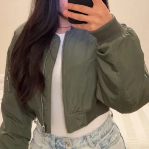 Grön croppad bomberjacka - Säljer en snygg croppad bomberjacka i mörkgrön färg med dragkedja framtill. Jackan har långa ärmar med breda muddar och en klassisk bomberkrage. Perfekt att styla med jeans eller kjol för en cool look.