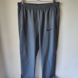 Grå träningsbyxor från Nike - Snygga grå träningsbyxor från Nike med elastisk midja och diskret svart logga på benet. Byxorna har raka ben och sidofickor, perfekta för sport eller chill. Materialet känns lätt och smidigt, vilket gör dem bekväma att bära hela dagen.