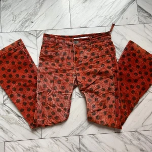 Orange manchesterbyxor med prickigt mönster - Unika orange manchesterbyxor med svarta prickar och bootcut-silhuett. Byxorna har klassiska fickor fram och bak, bälteshällor och dragkedja med knapp. Materialet är mjukt och ger en cool retrovibe. Perfekta för dig som vill sticka ut med färg och mönster.