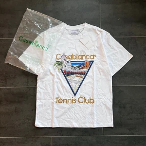 Casablanca Tennis Club - Vit t-shirt från Casablanca. Stort 'Casablanca Tennis Club'-tryck i guld och blått. Klassisk passform, rund hals och korta ärmar. Tillverkad i mjuk bomull.