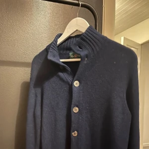 Mörkblå kofta från Alan Paine - Snygg mörkblå kofta från Alan Paine med ribbad hög krage och stora knappar framtill. Koftan har två fickor och är stickad i mjukt ullmaterial. Passformen är normal och ärmarna är långa, perfekt för kyliga dagar.