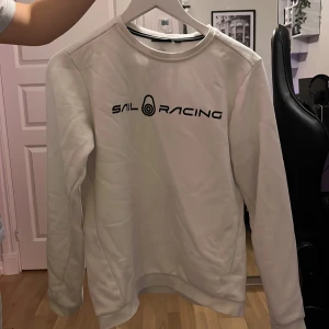 Vit Sail Racing sweatshirt - Vit sweatshirt från Sail Racing med svart logotyp tryckt på bröstet och en mindre logga i nacken. Tröjan har rund hals, lång ärm och är tillverkad i mjuk bomullsmix med insida i frotté. Passformen är normal och clean. Säljer då den inte passar mig längre.