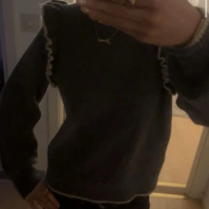 Mörkgrå stickad tröja med volang - Säljer en mörkgrå stickad tröja med ljusa volangdetaljer längs axlarna och kanten. Tröjan har långärmad passform och en lite loose siluett, perfekt för dig som gillar trendiga detaljer och vill ha något mysigt till garderoben.