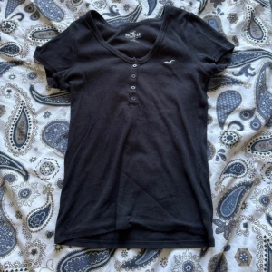 Marinblå t-shirt från Hollister - Svart t-shirt från Hollister med v-ringning och knappdetaljer framtill. Klassisk passform och korta ärmar, perfekt till jeans eller kjol. Liten vit logga broderad på bröstet. Mjuk och skön bomullskvalitet.
