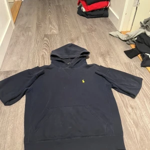 Mörkblå hoodie från Polo Ralph Lauren - Mörkblå hoodie från Polo Ralph Lauren med korta ärmar och klassisk huva. Tröjan har en gul broderad logga på bröstet och en stor magficka framtill. Tillverkad i mjuk bomull, perfekt för en chill och avslappnad stil.