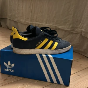 Adidas Gazelle blå/gula sneakers frakt betalas själv - Säljer ett par Adidas Gazelle sneakers i mörkblå mocka med gula detaljer och klassiska tre ränder på sidan. Skorna har snörning, vit platt sula och Arsenal-logga på plösen. Perfekt för dig som vill sticka ut med färg och stil. Kommer med extra snören i färgen gul. Pris går att diskutera.