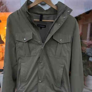 Olivgrön vindjacka från J.Lindeberg - Snygg olivgrön vindjacka från J.Lindeberg med hög krage, dragkedja och tryckknappar framtill. Jackan har två bröstfickor med lock och två sidofickor, samt en diskret svart logotyp på ärmen. Perfekt för dig som gillar stilren och funktionell design. Jackan är knappt använd och i väldigt gott skick!