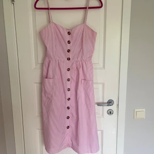 Rosa klänning - Söt rosa- och vitrandig klänning med knappar framtill och smala axelband. Klänningen har två fickor och smockad rygg❤️